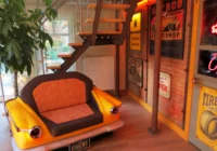 Gelbes Cadillac-Hecksofa mit Rückleuchten vor einer Retro-Werkstattfassade